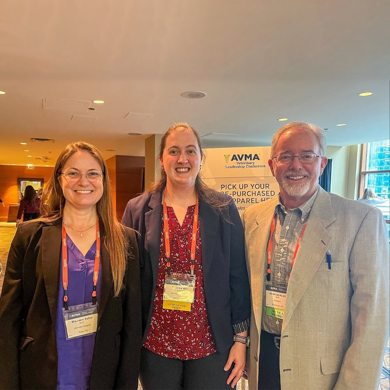 Three people, two women and one man, stand indoors at a conference, smiling at the camera. They wear business attire and conference badges. A sign in the background reads “AVMA Leadership Conference.”.