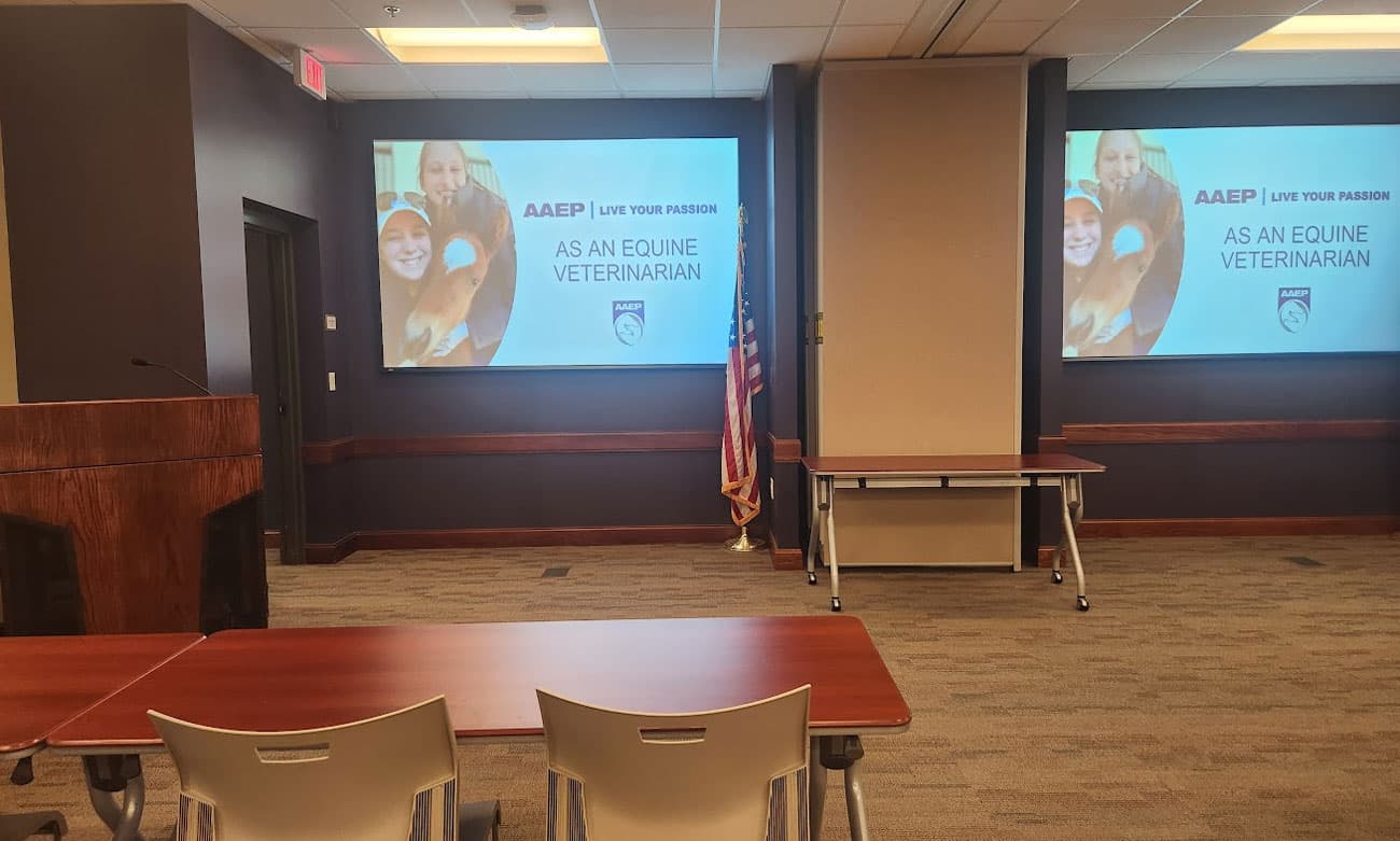 A conference room with a podium, chairs, tables, and two screens displaying "AAEP Live Your Passion: As an Equine Veterinarian" with images of smiling people and a horse. An American flag stands near the center.