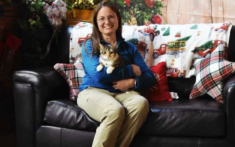 A woman sits on a dark sofa, smiling and holding a cat. The background features Christmas decorations, a festive blanket with red trucks, and plaid pillows, creating a cozy holiday atmosphere.