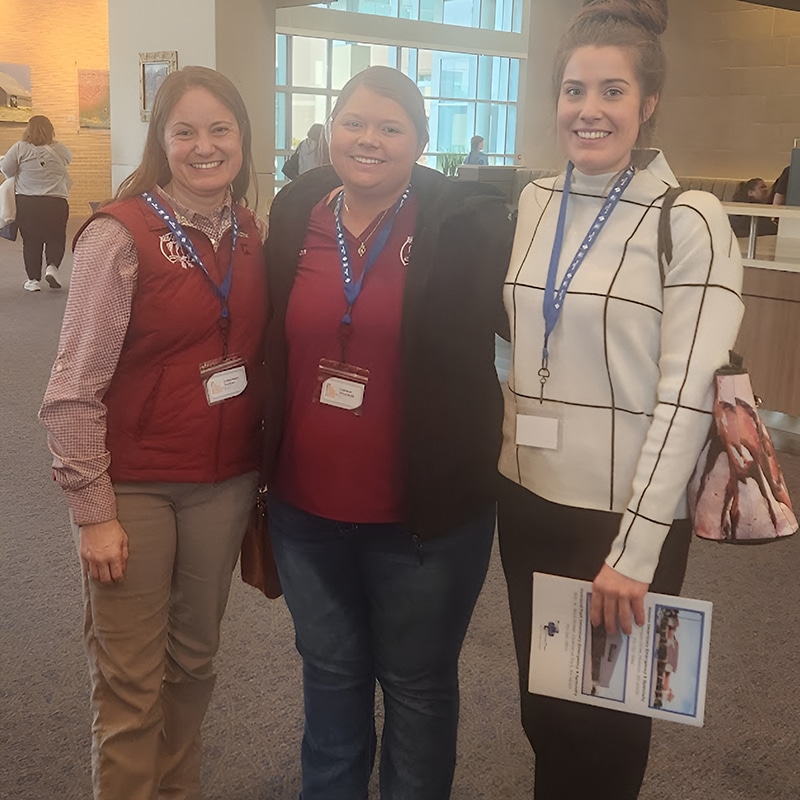 Three women stand indoors, smiling at the camera. They wear name badges and lanyards; two are in maroon shirts, and one wears a white sweater with a black grid pattern, holding papers and a patterned bag.