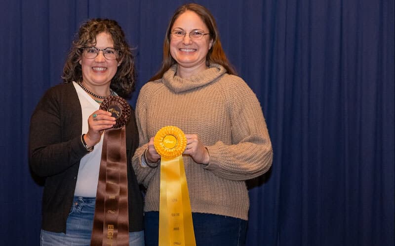 Two women stand in front of a blue curtain, smiling and holding award ribbons—one brown and one yellow. Both are wearing glasses and sweaters, celebrating their achievements.