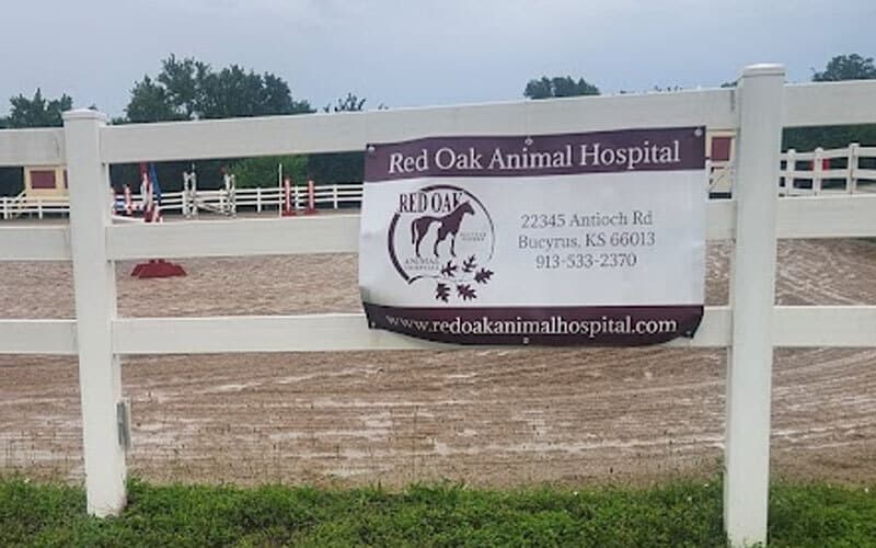 A banner for Red Oak Animal Hospital hangs on a white fence near a dirt arena, displaying the clinic's address, phone number, website, and a logo with two animal silhouettes.