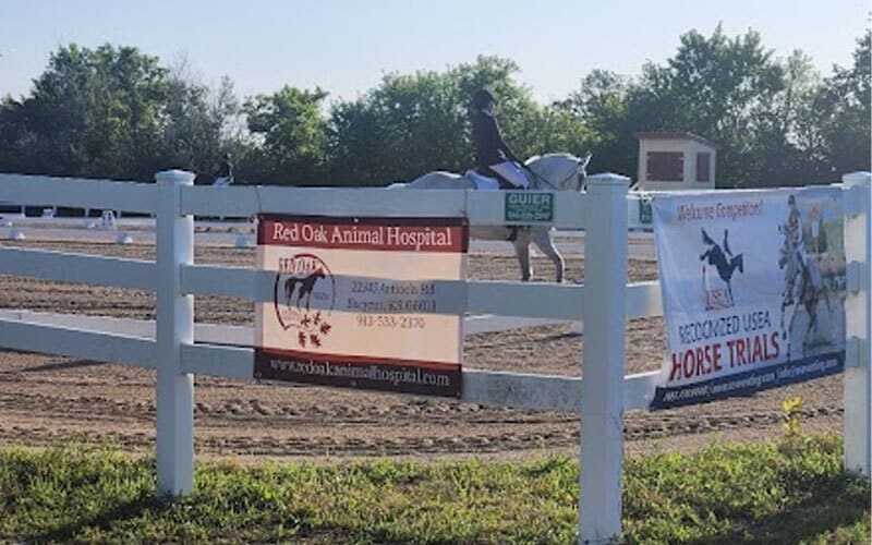 A person riding a white horse in an outdoor arena, with two banners on a white fence in the foreground advertising Red Oak Animal Hospital and a recognized USA horse trials event. Trees and a small building are in the background.