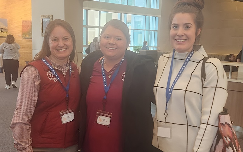 Three women wearing conference badges and blue lanyards pose together indoors. Two wear maroon shirts and vests, and one wears a white checkered sweater. Other people and large windows appear in the background.