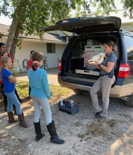 A woman in uniform shows a large animal skull to a group of children standing next to an SUV with its trunk open. The setting appears to be outdoors near a building and some trees.