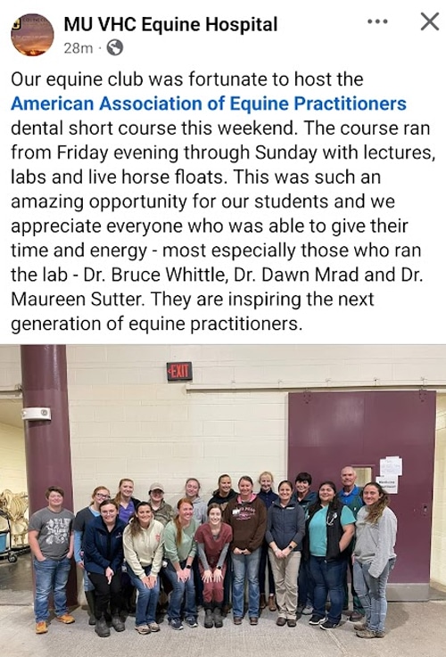A group of about 18 people, mostly in casual clothes, stand smiling in a row indoors against a plain wall. They are posing together, some with arms around each other, after an equine practitioners event.