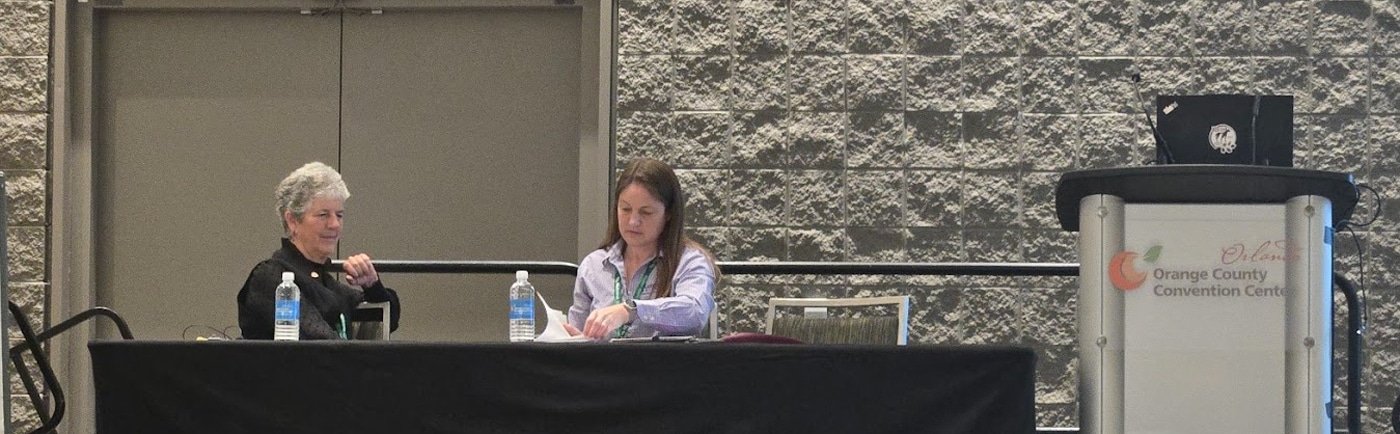 Two women sit at a table on a stage with water bottles, papers, and microphones. A podium labeled “Orange County Convention Center” stands nearby. The background features a textured gray wall.
