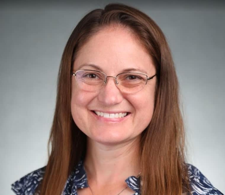 A woman with long brown hair, glasses, and a blue patterned top smiles at the camera against a plain grey background.