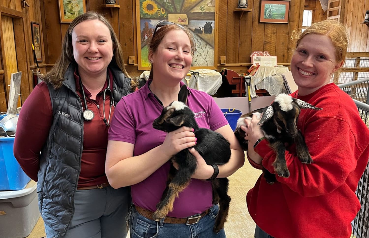 Three smiling women hold baby goats indoors. The women wear casual work clothes, including vests and polos, with stethoscopes around their necks. The background features wooden walls, paintings, and farm equipment.