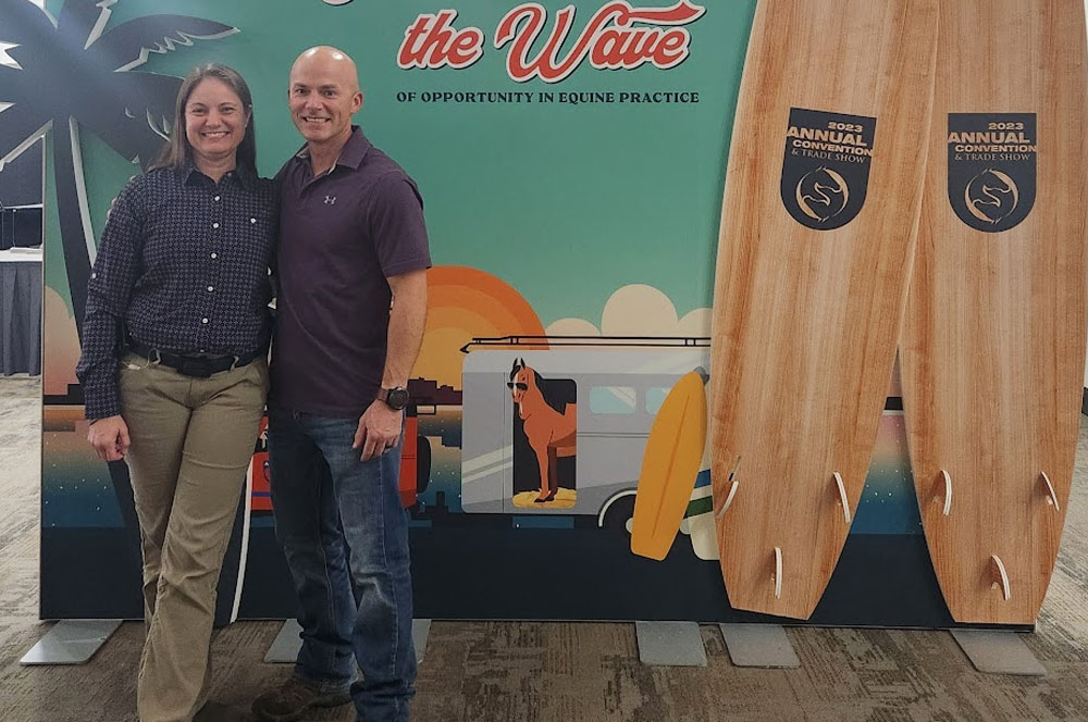 Two people stand smiling in front of a colorful backdrop featuring a beach scene, a van, a dog, palm trees, and two large surfboards labeled "2023 Annual Convention & Trade Show.
