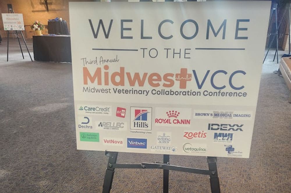 A large sign welcomes attendees to the Third Annual Midwest Veterinary Collaboration Conference (Midwest VCC), displaying logos of various sponsors in a conference venue setting.