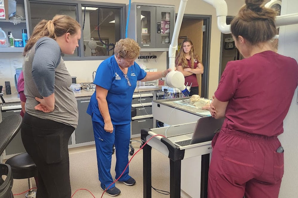 A group of women in scrubs and casual wear observe a woman in blue scrubs operating veterinary equipment over a small animal model in a clinical lab setting. Shelves with supplies and cabinets line the background.