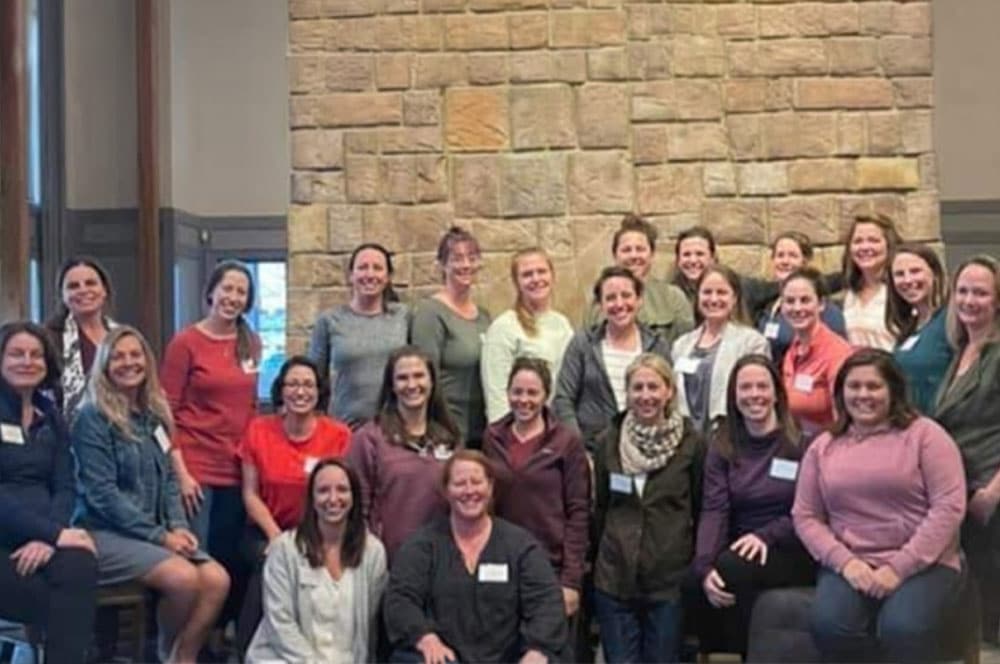 A group of 24 women pose together indoors in front of a stone wall, smiling at the camera. Most are standing, while a few are seated in front. They wear casual clothing and name tags.