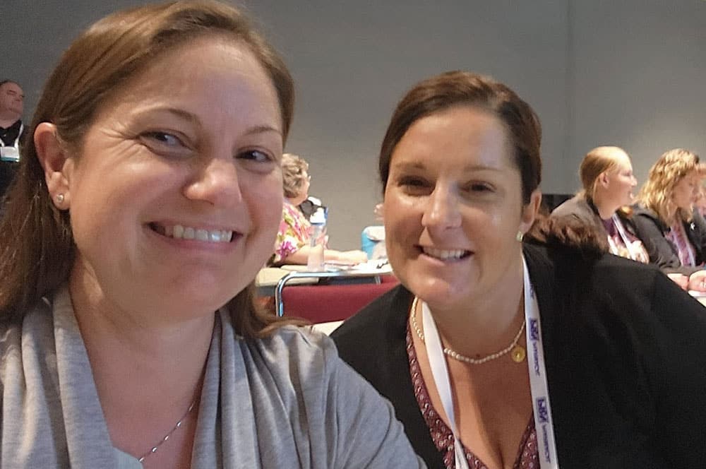 Two women smiling and posing for a selfie at a conference or seminar, with other attendees visible in the background seated at tables.