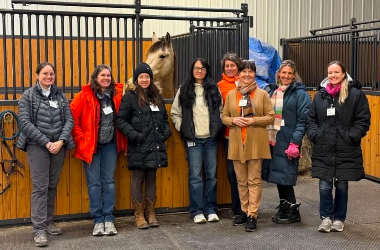 A group of eight women in winter clothing stand smiling together in front of a horse in a stable with wooden and metal stalls.