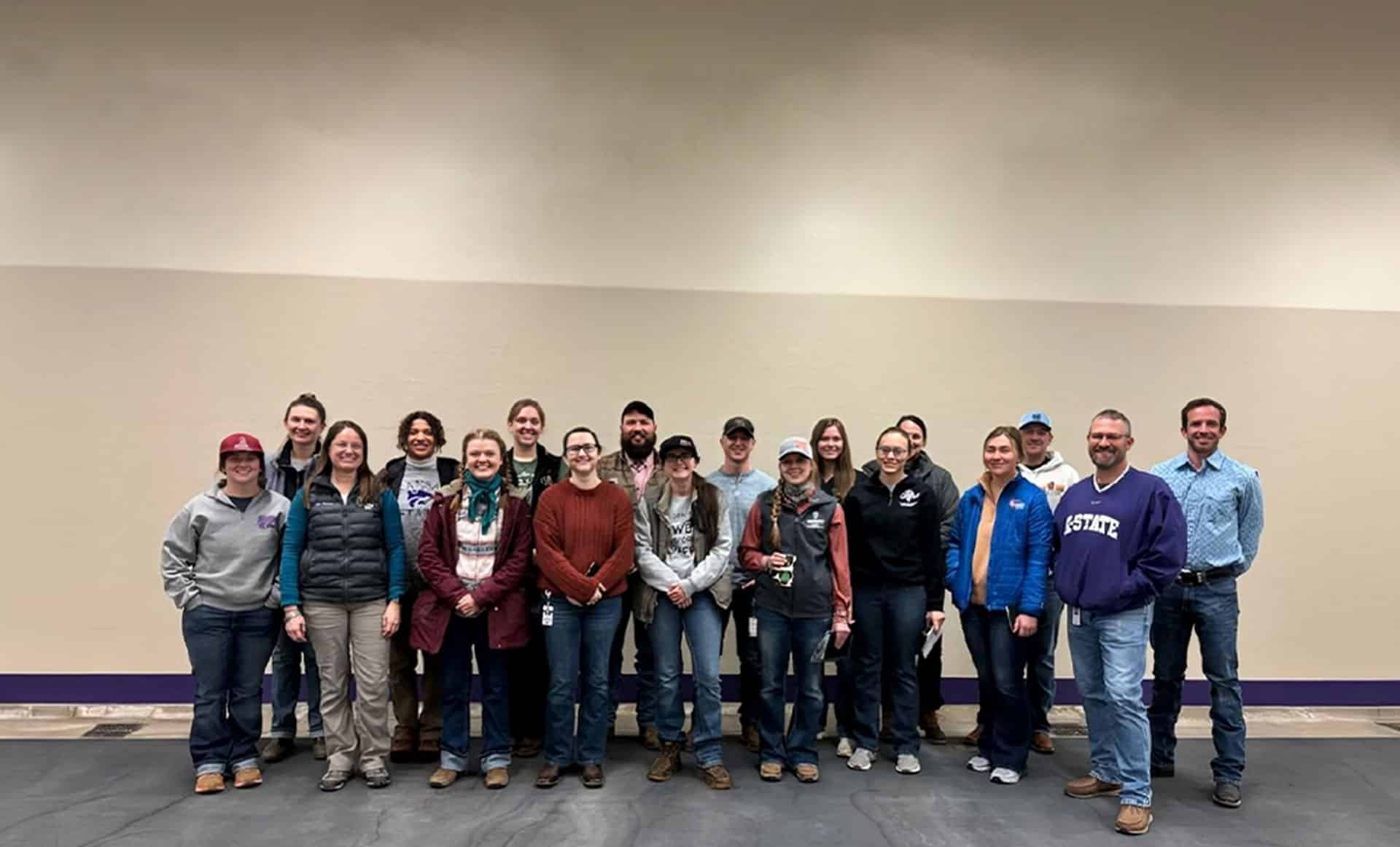 A group of 19 people, dressed casually in jackets, jeans, and sweaters, stand indoors in a single line against a beige wall, smiling at the camera.